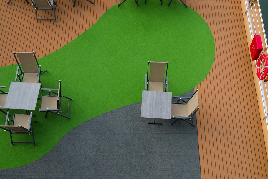 An aerial view of an outdoor deck with tan flooring, a curved green turf section, tables, and chairs.