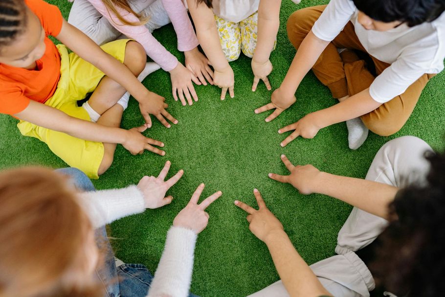 Several people sit in a circle on green turf, each pointing two fingers toward the center.