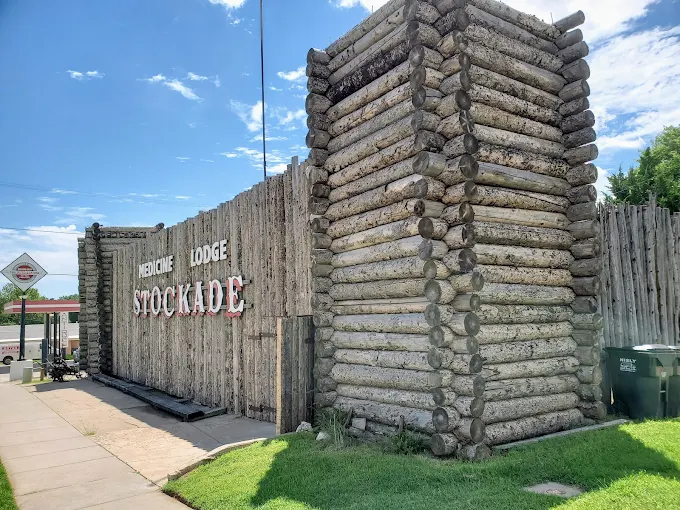 Wooden lodge sign reading “Stockade” beside a rustic log-fence entrance under a blue sky