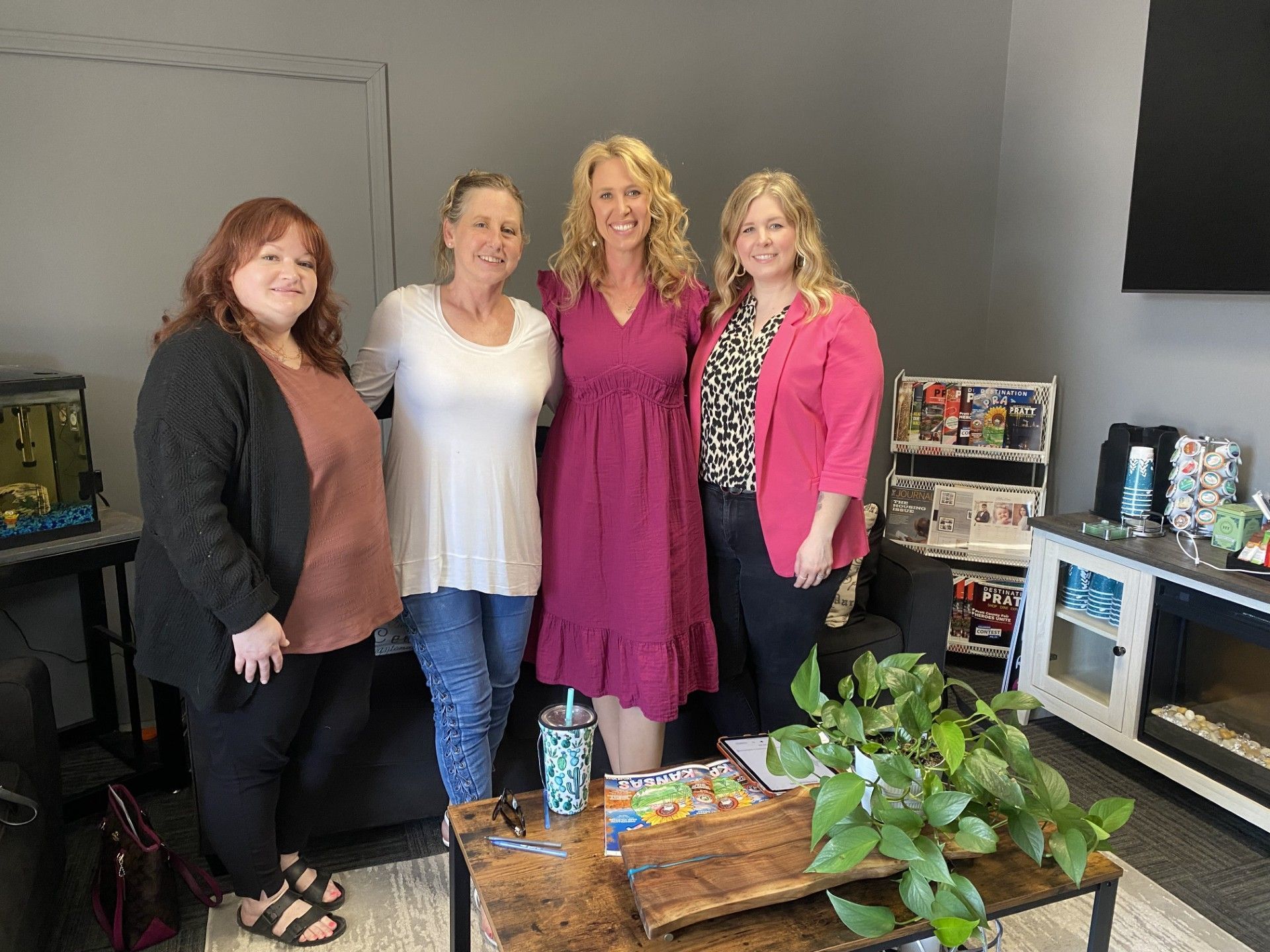 Four women standing together in an office lounge beside a coffee table and display shelves