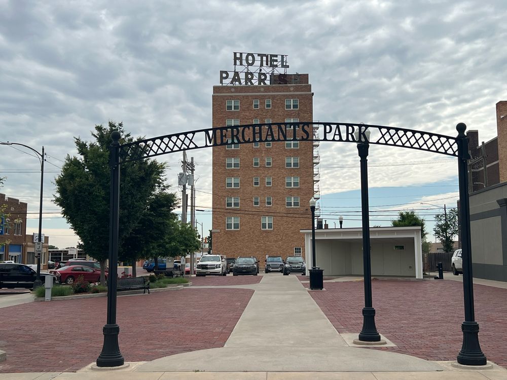 Brick entrance arch marked “Merchants Park” with a hotel building behind it under a cloudy sky
