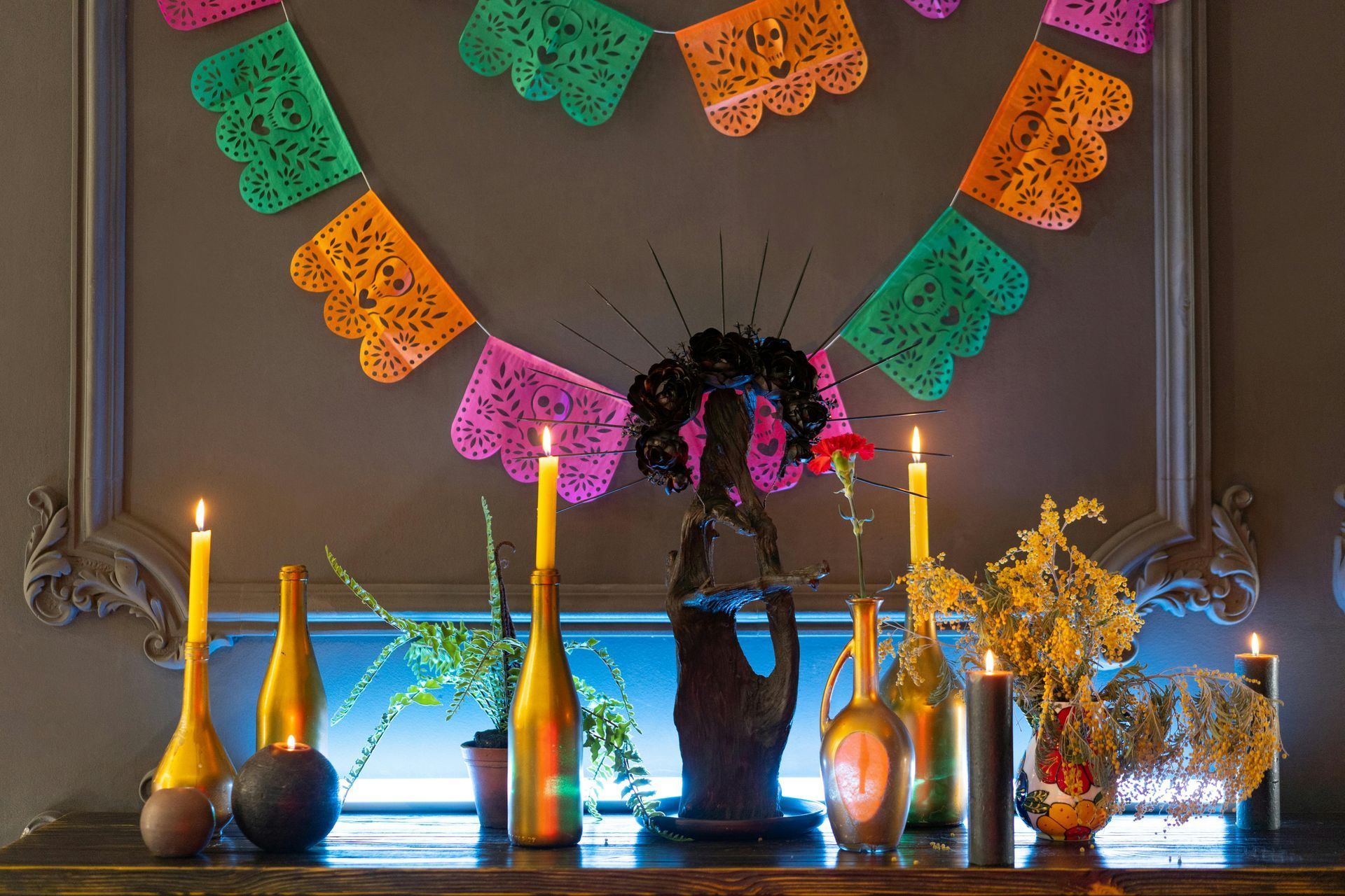 Decorated altar with candles, bottles, flowers, and colorful papel picado garland against a framed wall