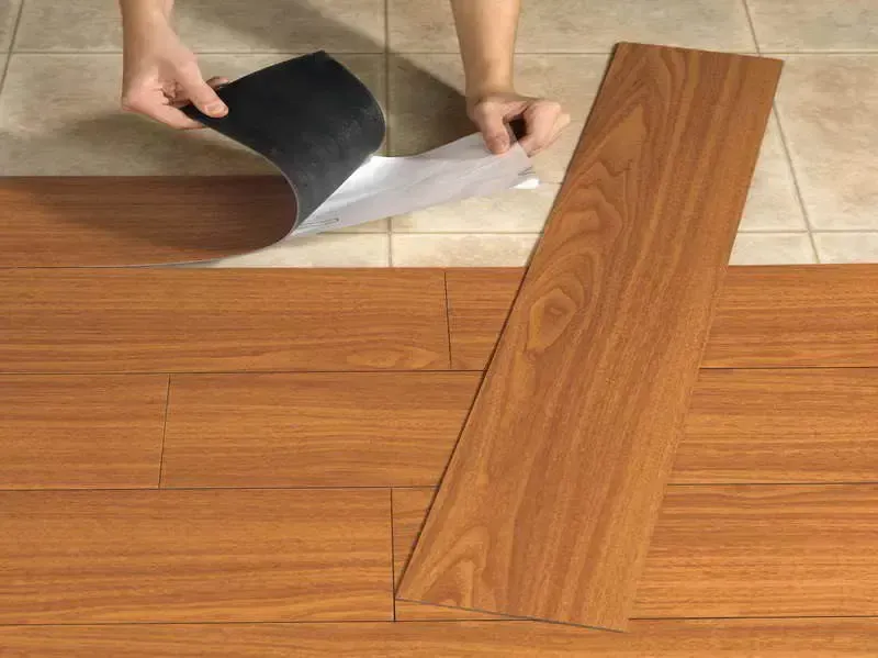 Person installing peel-and-stick wood-look flooring. Hands peeling backing from a plank, over existing tile floor.