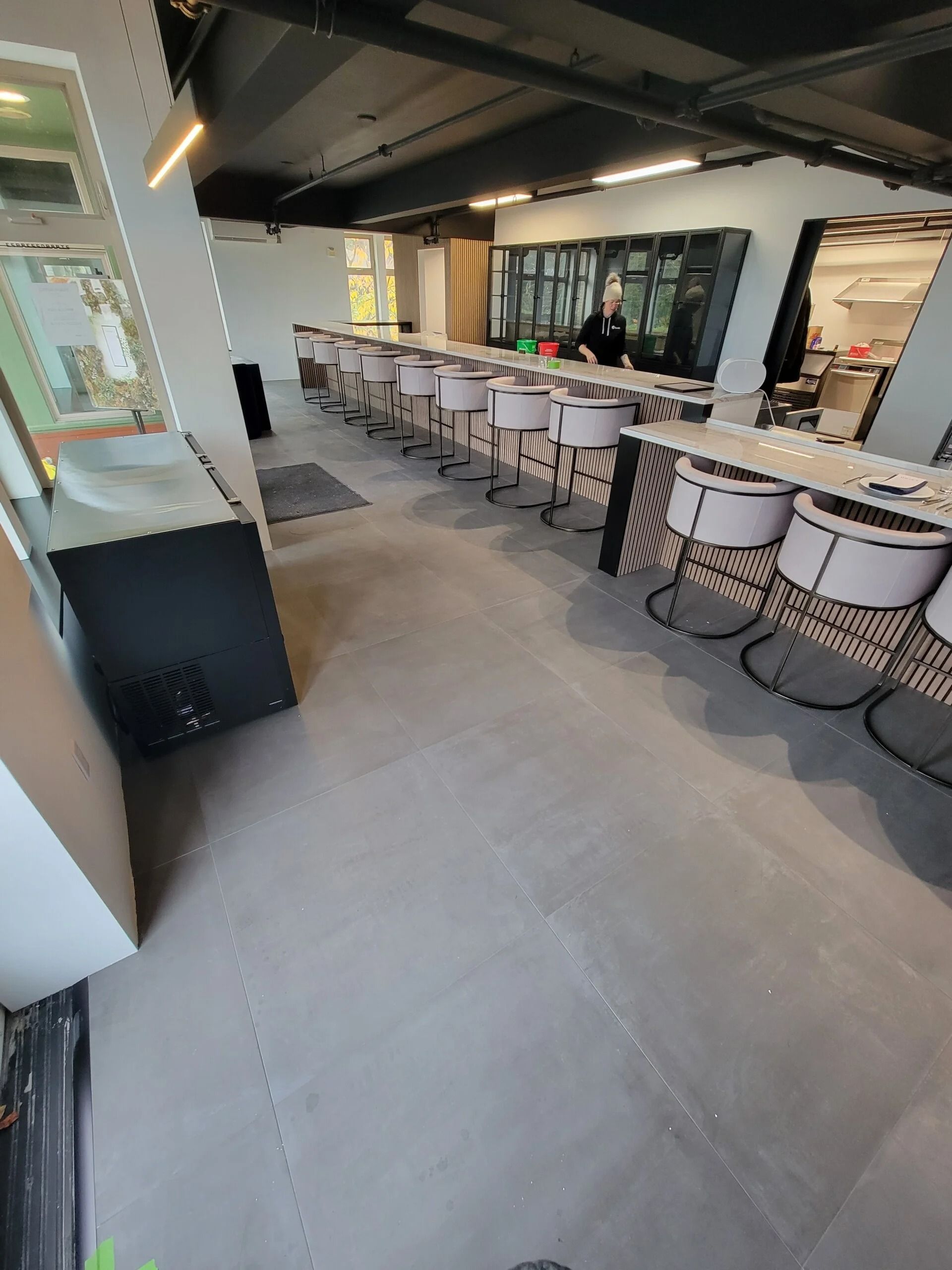 Long, narrow room with a counter and chairs. A person stands behind the counter, room has gray floors and a black ceiling.
