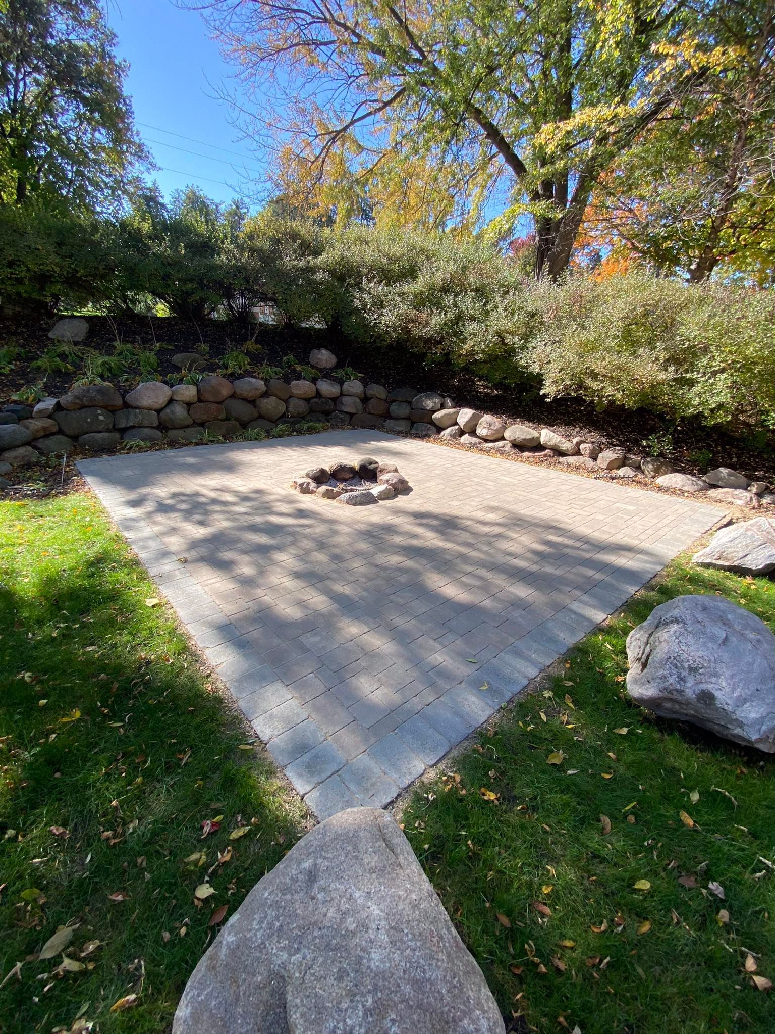 Image of an amazing lanscape design project in Wayzata, MN. You can see a custom built fire pit surrounded by pavers and a rock retaing wall. Above the retaining wall you can see variegated privet shrubs planted right above the retaining wall.  