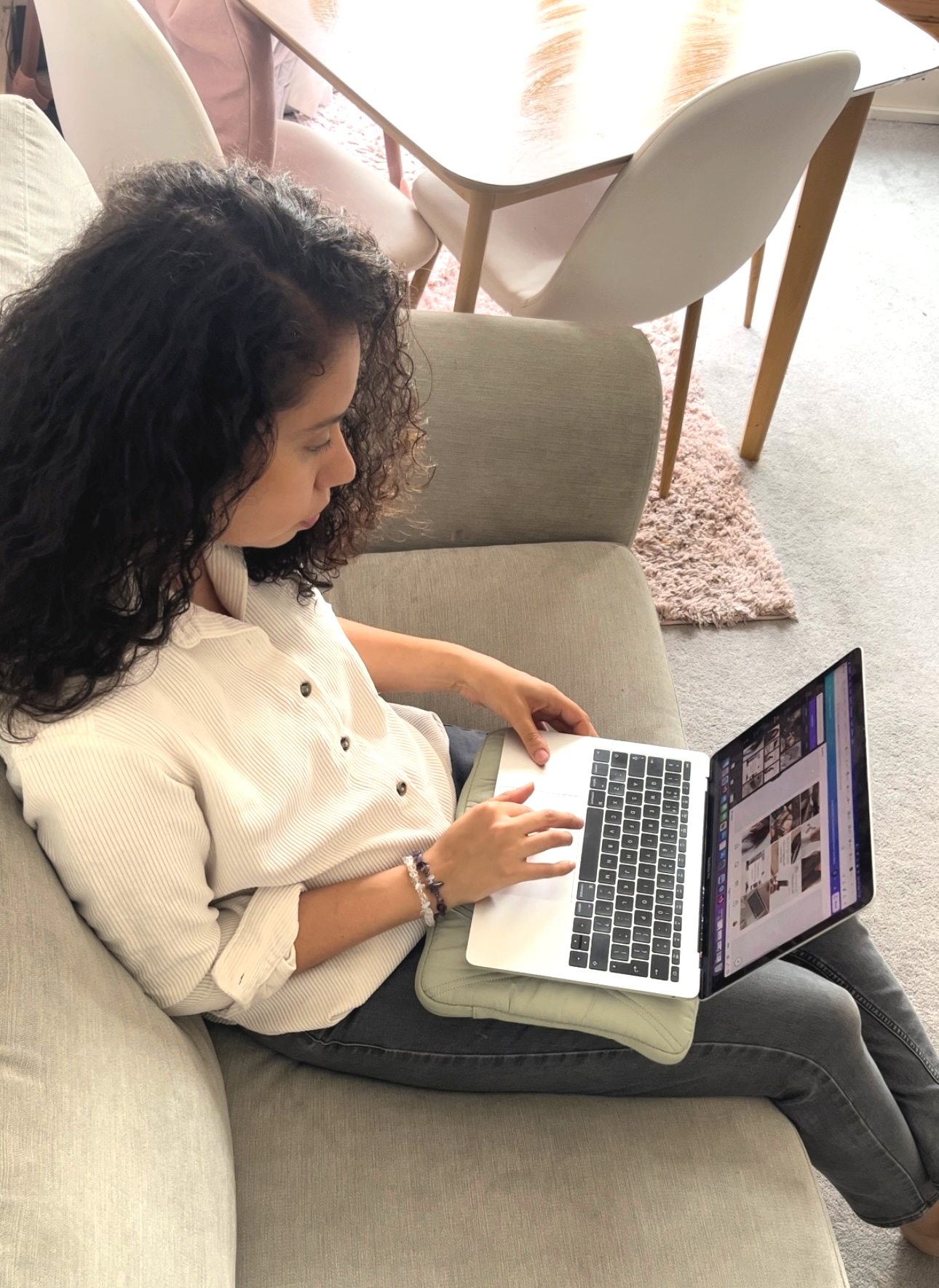 Woman with curly hair sits on a bench outdoors, working on a laptop in the sun.