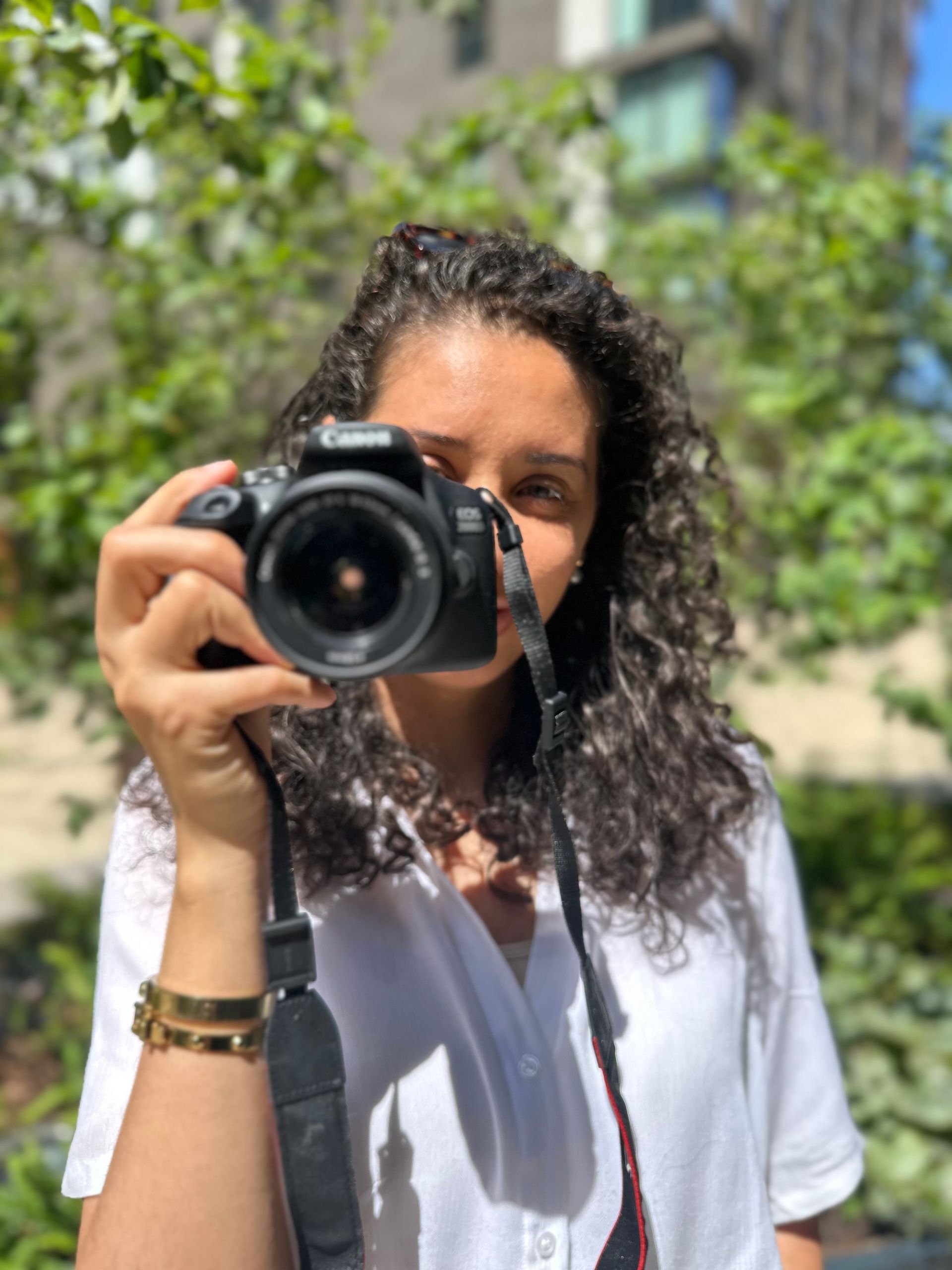 Woman with curly hair holding a camera, partially obscuring her face. Outdoors with greenery in the background.