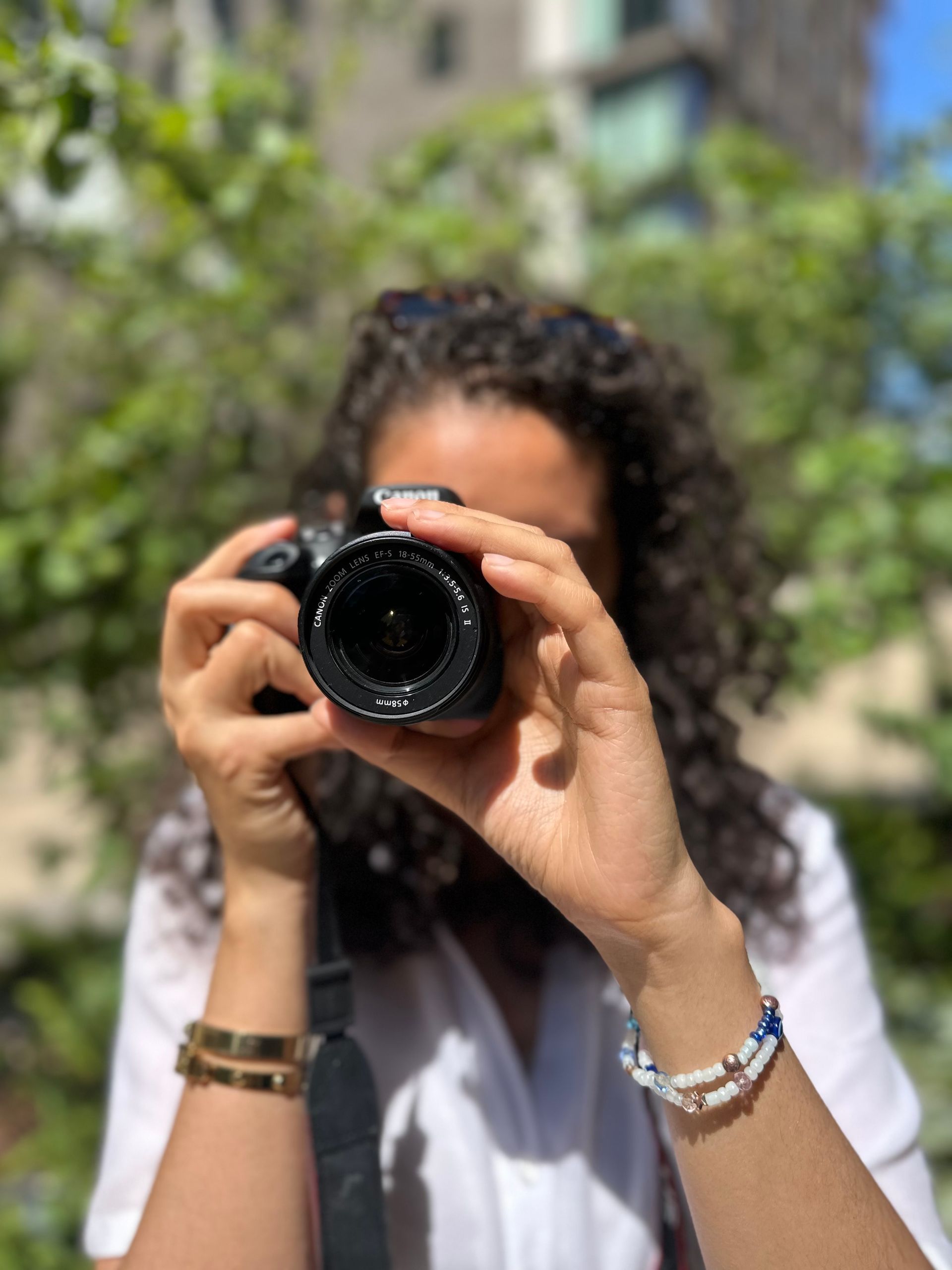 Woman with curly hair taking a photo with a camera, hands in focus, outdoors.