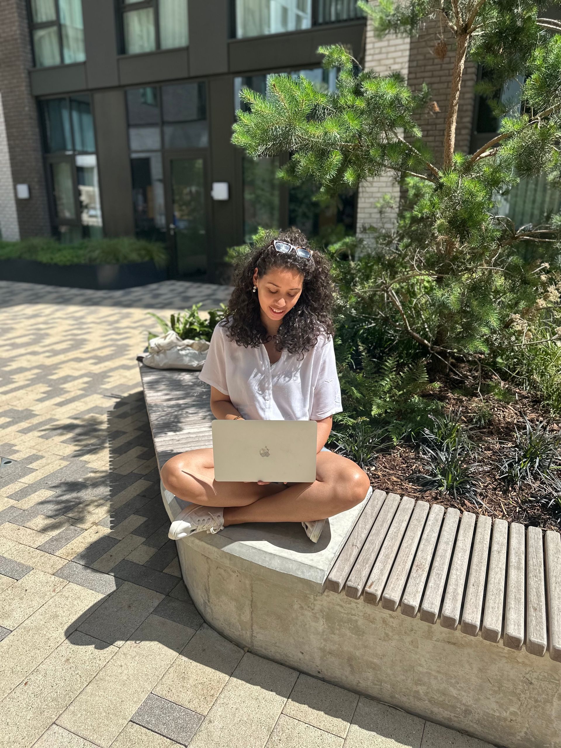 Woman sitting cross-legged, using laptop outdoors on a sunny day, in front of a building.