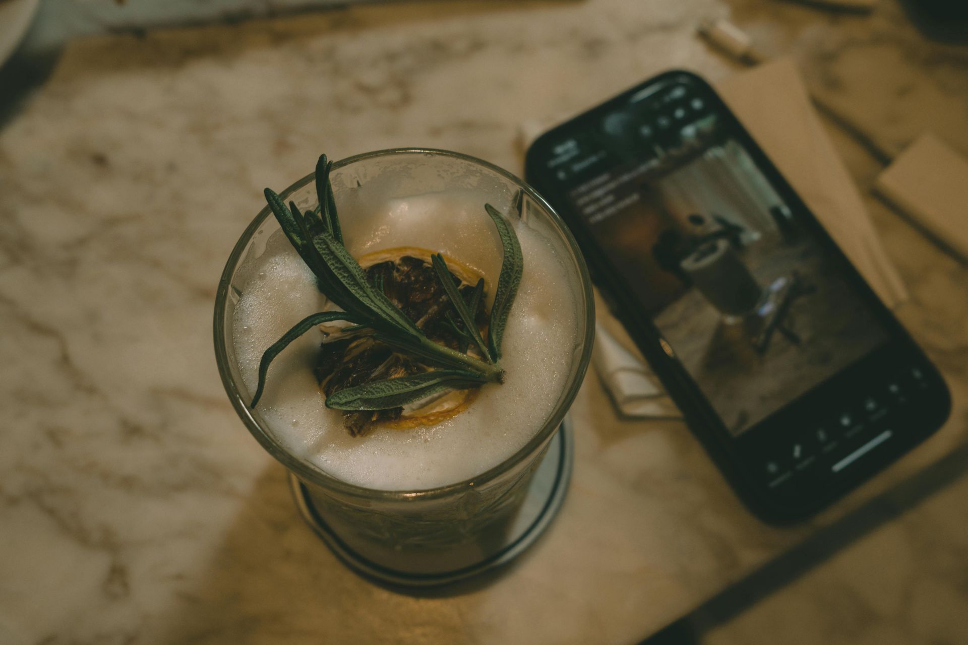 Cocktail with rosemary garnish and a phone displaying a photo, resting on a marble table.