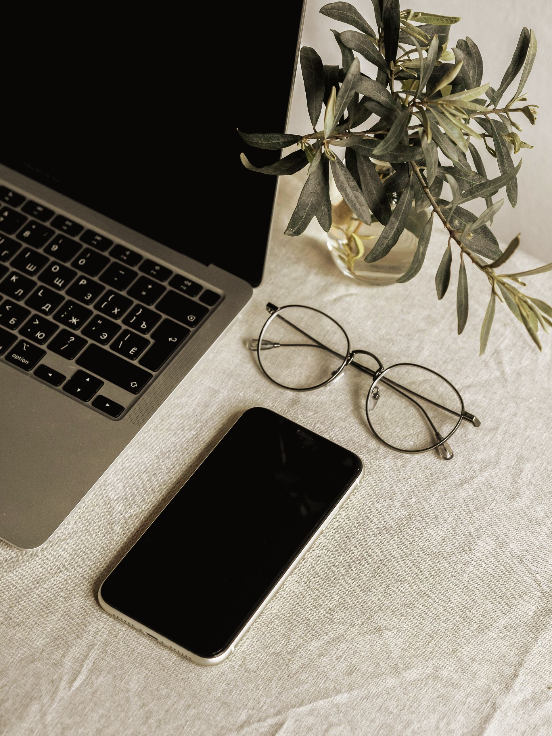Laptop, phone, and eyeglasses on a white tablecloth with a vase of green sprigs, neutral colors.