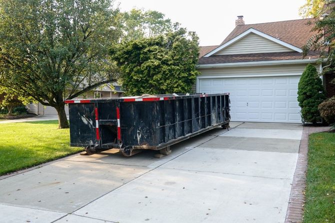 Black construction dumpster in a driveway beside a garage and tree-lined suburban house.