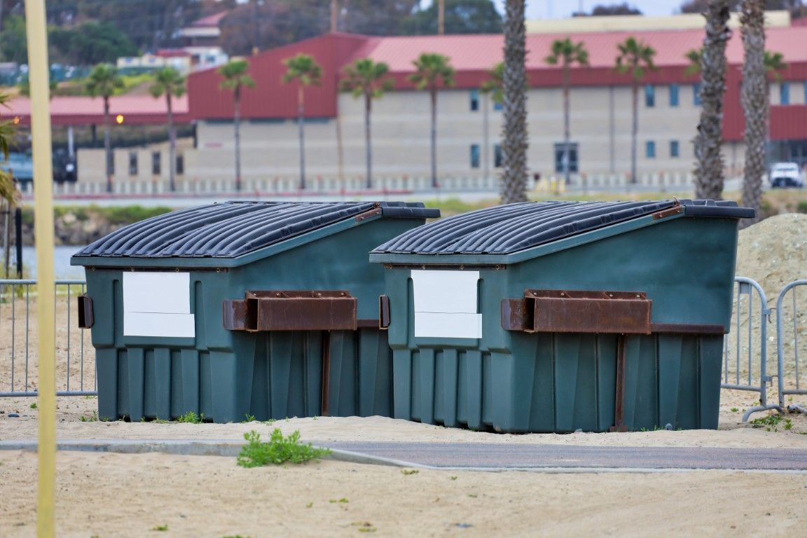 Two green dumpsters with black lids beside a sandy path, with buildings in the background.