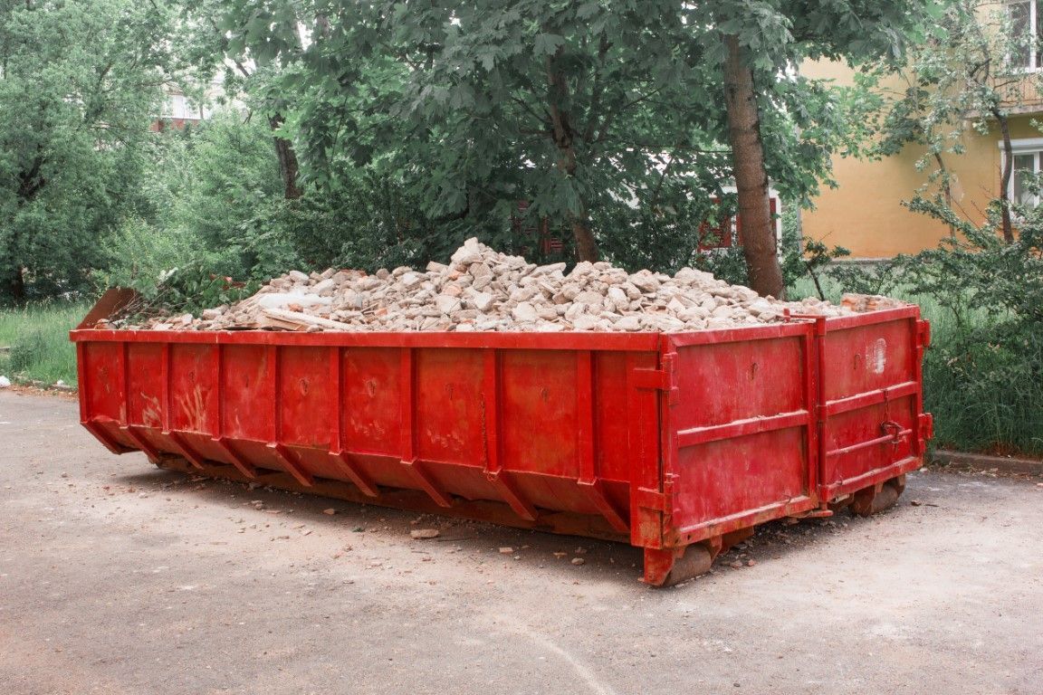 Red construction dumpster filled with rubble on a dirt lot, with trees in the background
