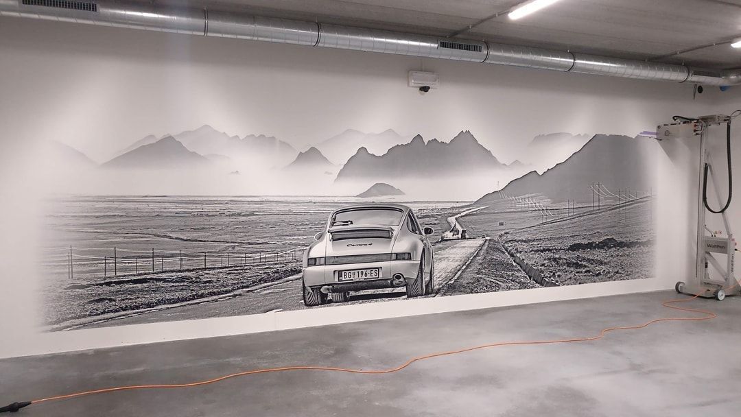 Zwart-wit muurschildering van een auto op een weg met een berglandschap.