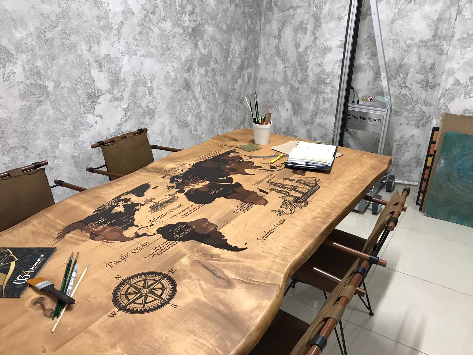 Houten tafel met een wereldkaart, penselen en stoelen in een kamer met een grijze muur.