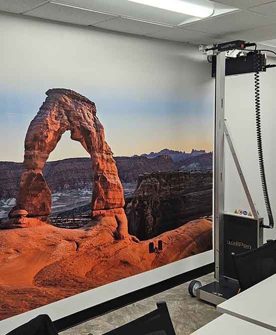 Een grote robot op wielen tegen een muurschildering van Delicate Arch in een felverlicht kantoor.