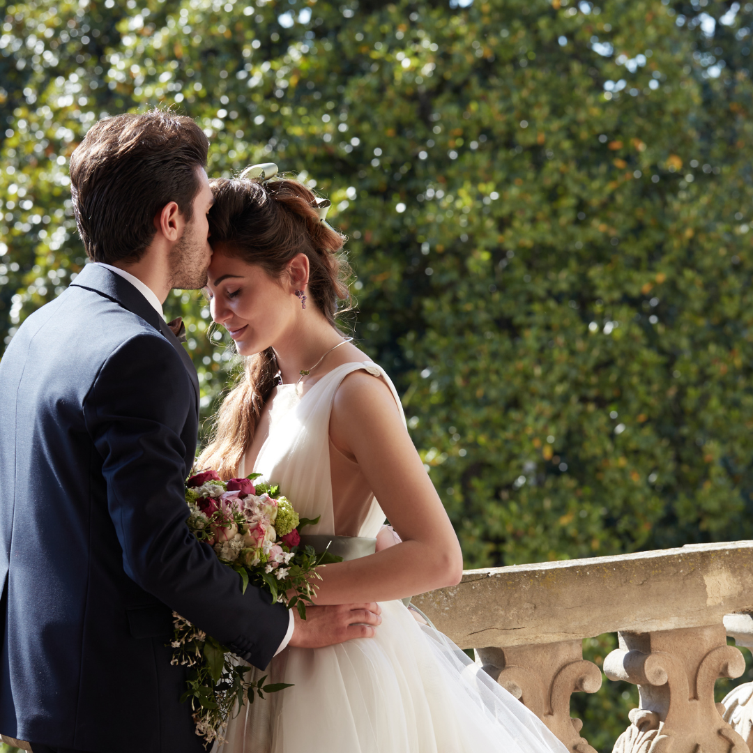 Groom kisses bride's forehead on a balcony. She wears a white dress, holding a bouquet. Sunny day.