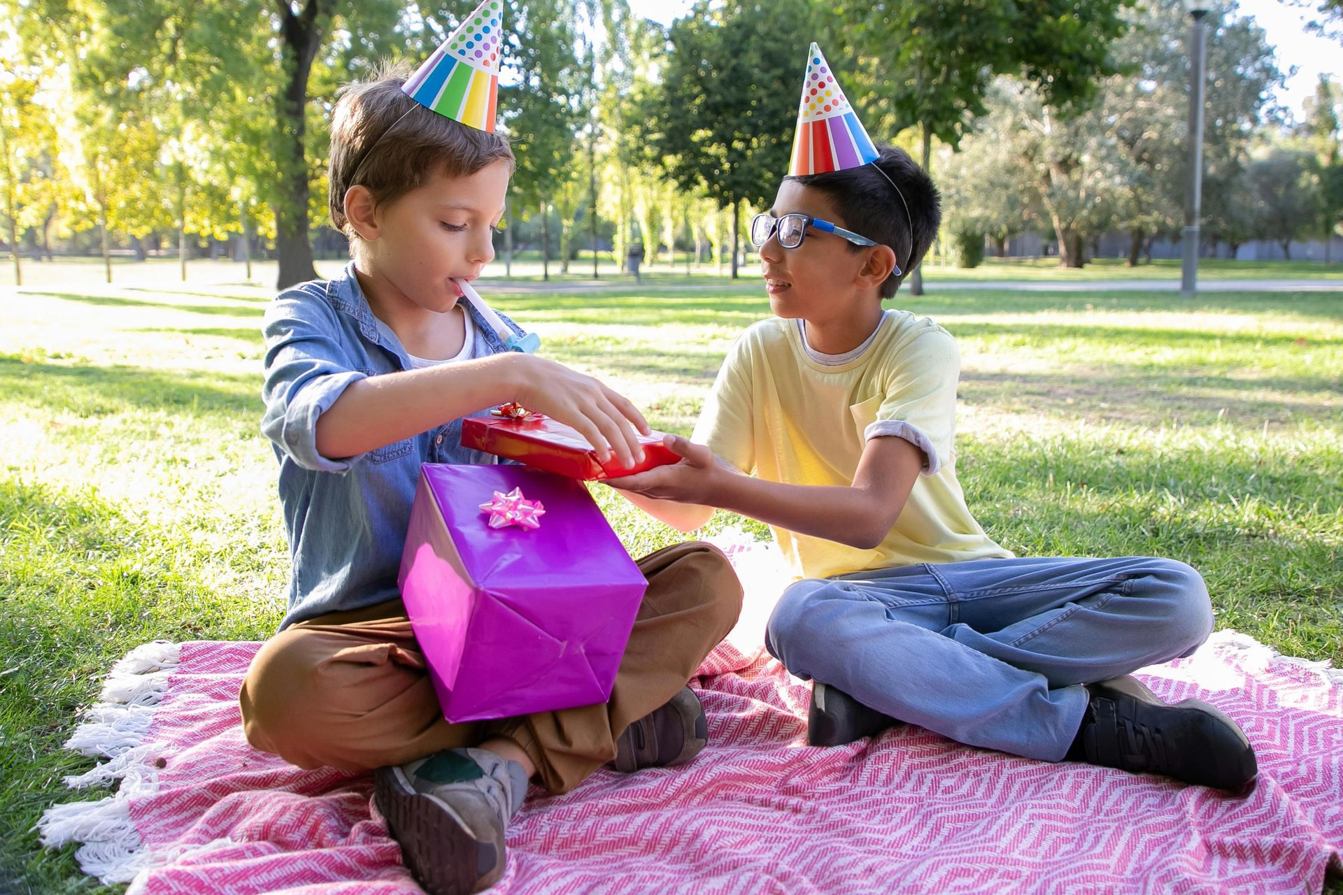 Two boys in party hats open gifts on a blanket in a park.