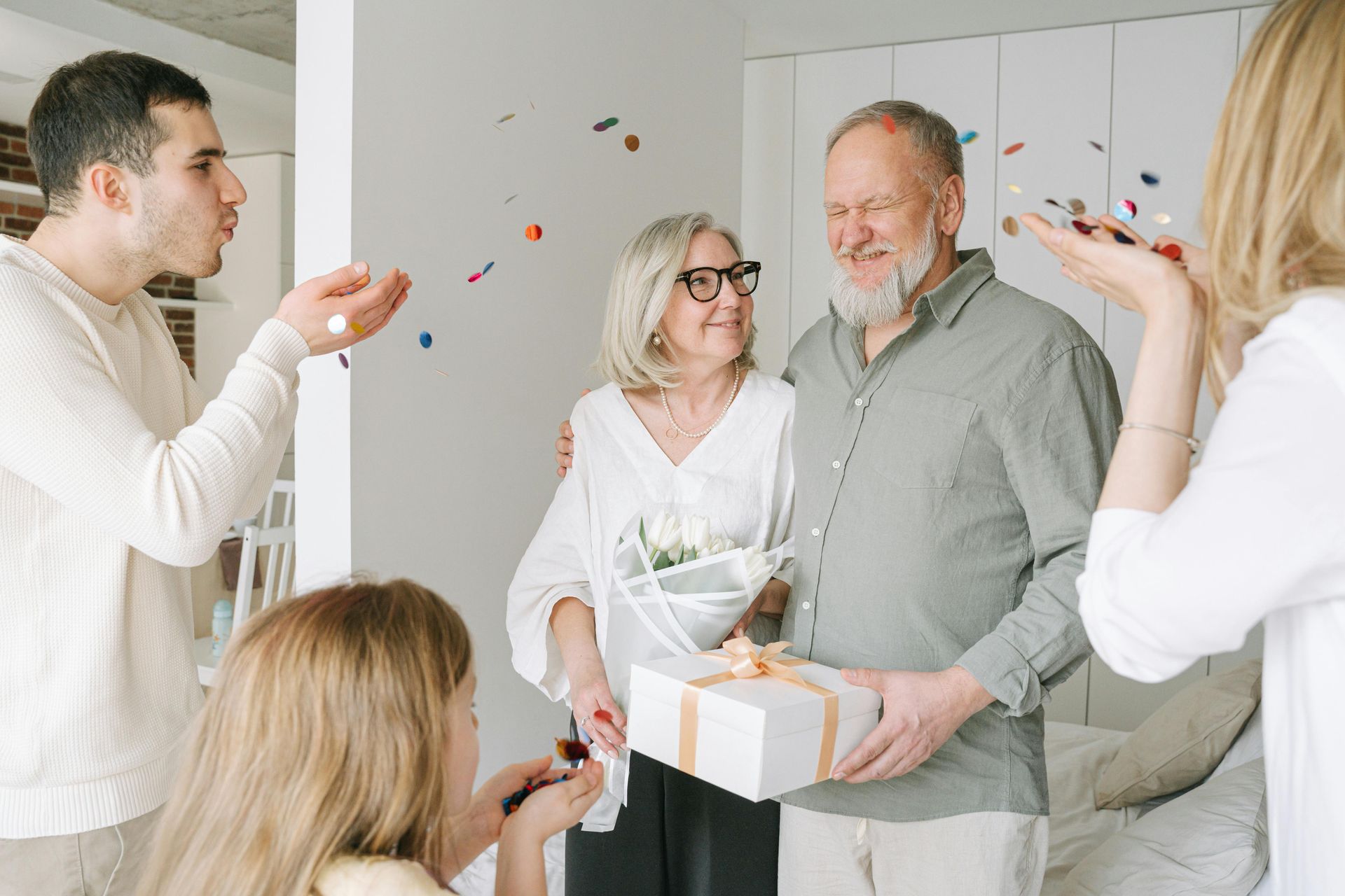 Family celebrates with confetti; elderly couple embraces, holding a gift and flowers.
