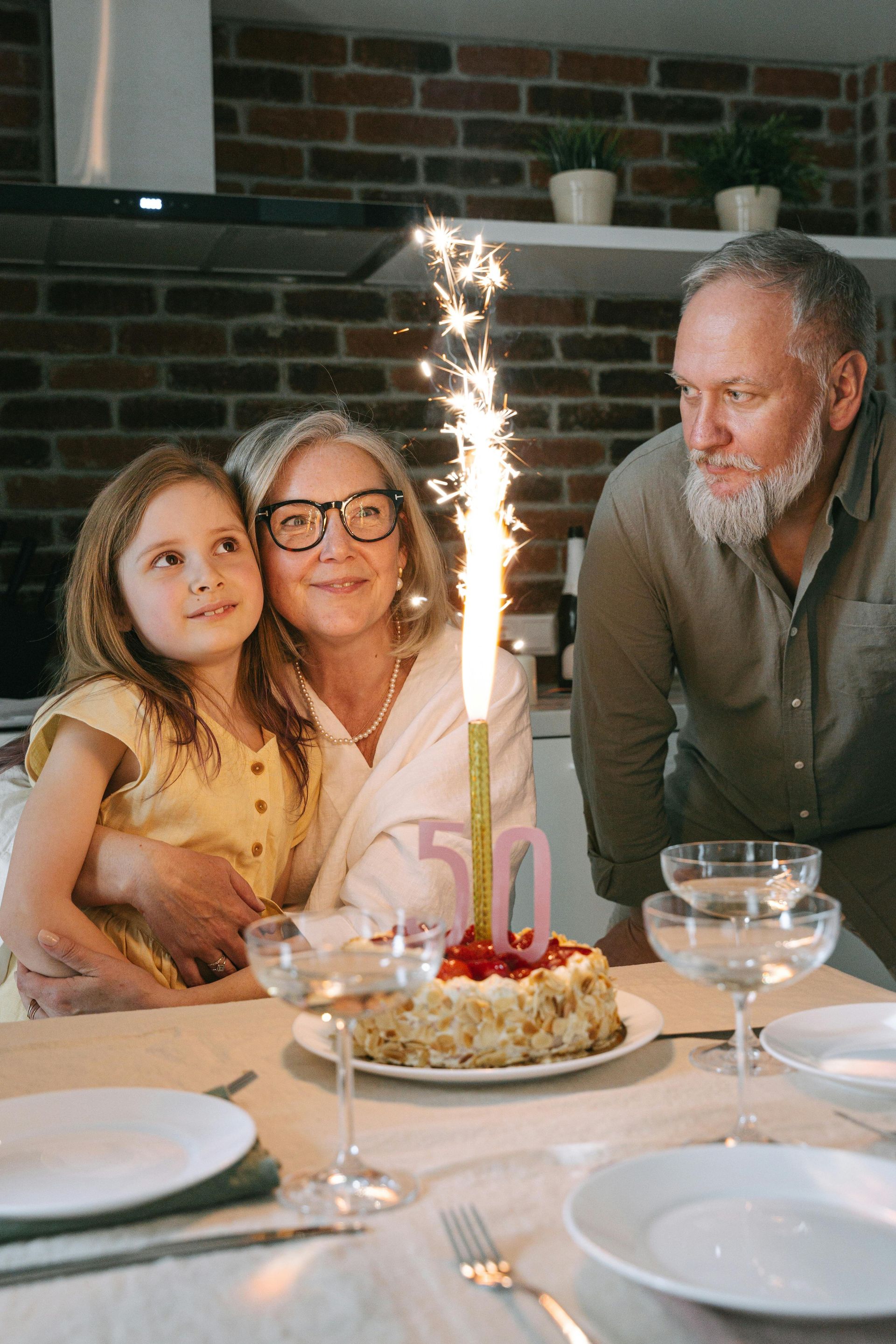 Woman with glasses hugs a child, watching a cake with a sparkler. A man looks on. Kitchen setting.