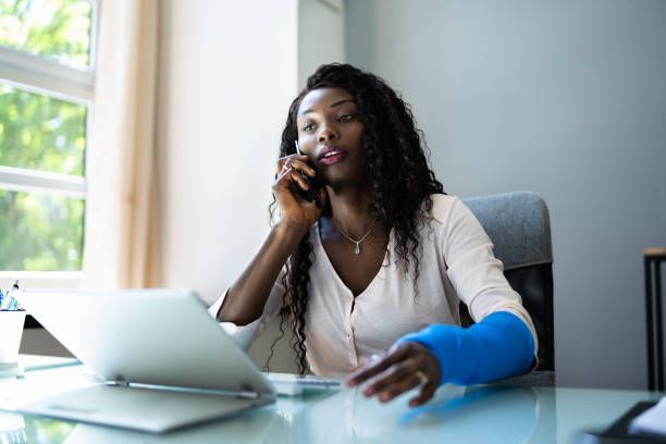 A woman with a cast on her arm is sitting at a desk talking on a cell phone.