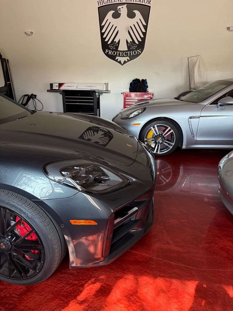 Dark grey sports car and silver Porsche in a garage with a red floor, under a crest.