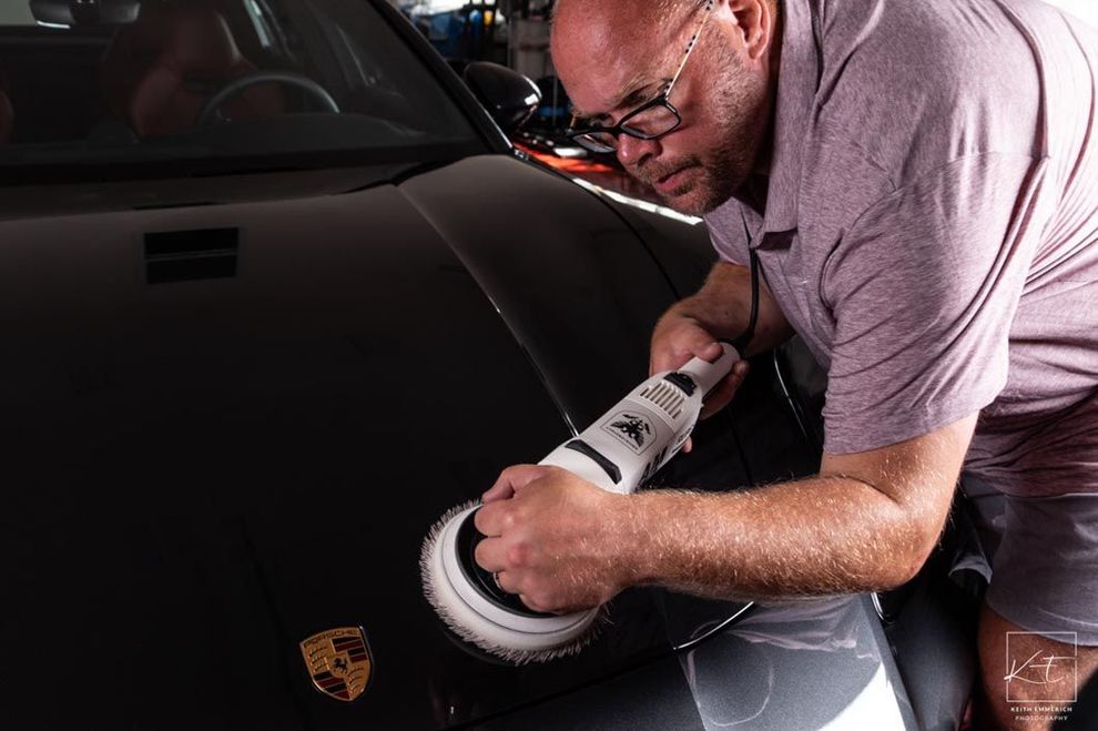 Man polishing a black car's hood with a machine, in a garage.