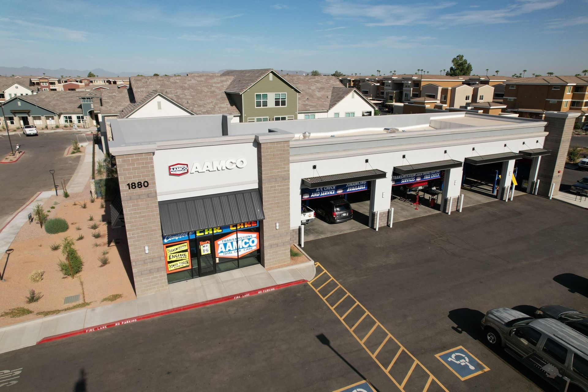 Exterior view of an AAMCO auto repair shop with open service bays, in front of a residential area under a blue sky.