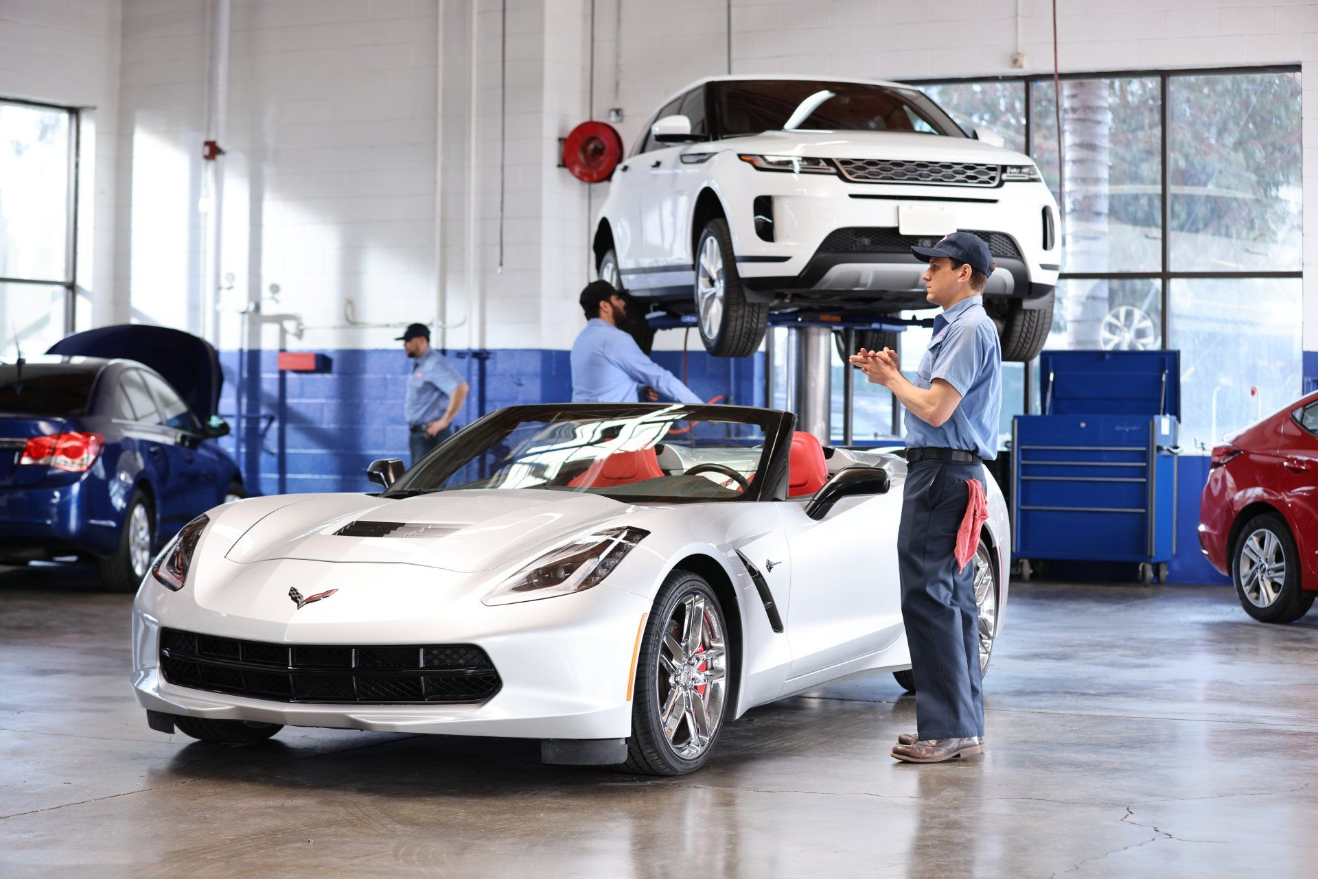 White sports car in AAMCO auto shop with a lifted SUV. Mechanics working, tools visible.