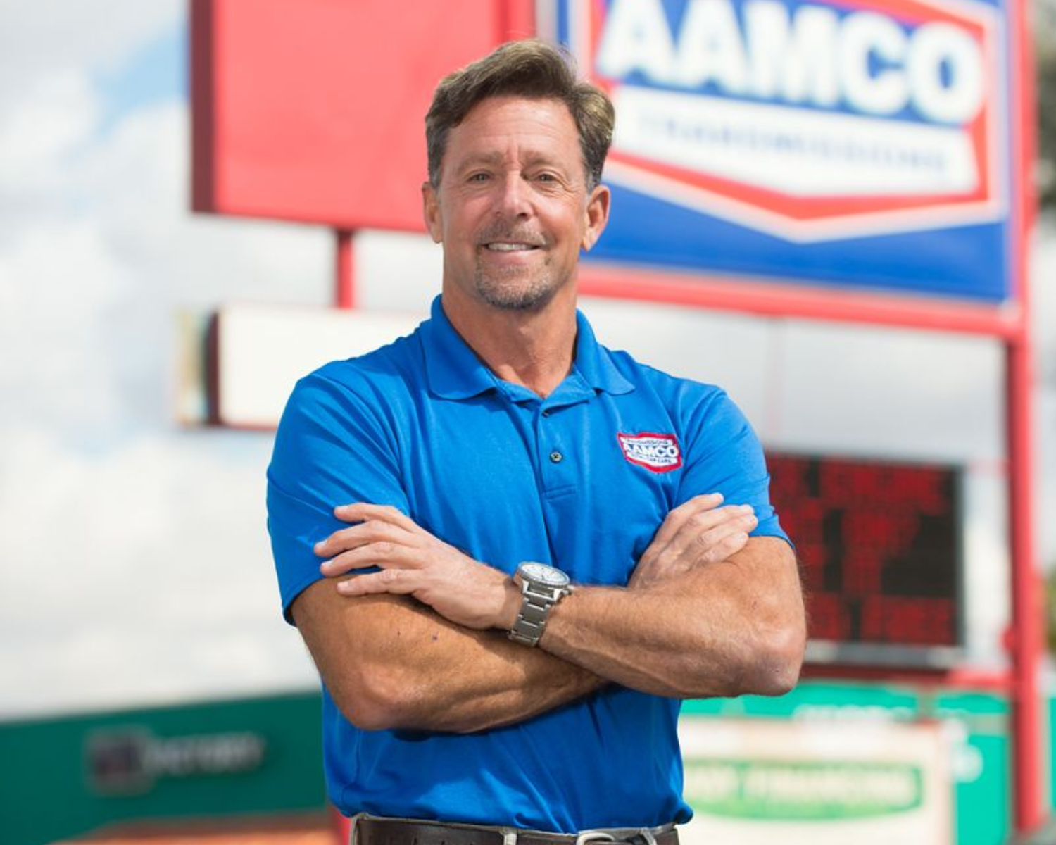A man in a blue polo shirt with arms crossed, smiling, standing in front of an AAMCO sign.