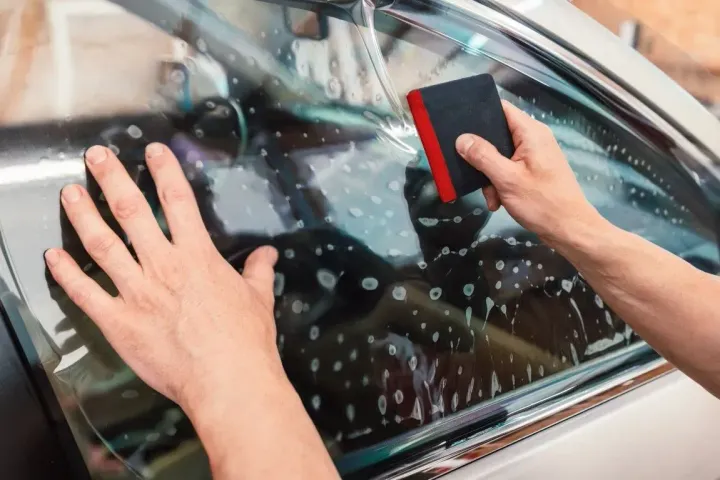 Hands cleaning a car window with a black sponge and red squeegee, water droplets visible on the glass