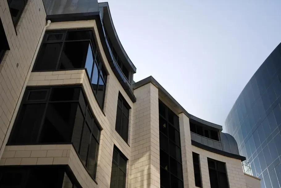 Curved stone office building beside a reflective glass tower against a bright sky