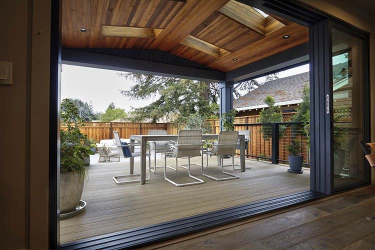 Sliding glass doors open onto a deck with dining set, beneath a wooden ceiling with skylights.