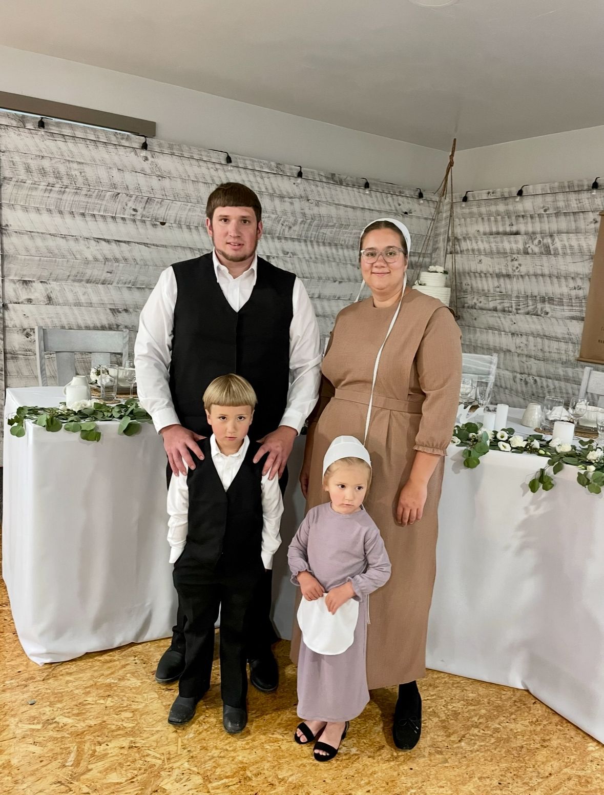 Amish family posing by decorated table: man, woman, and two young children.