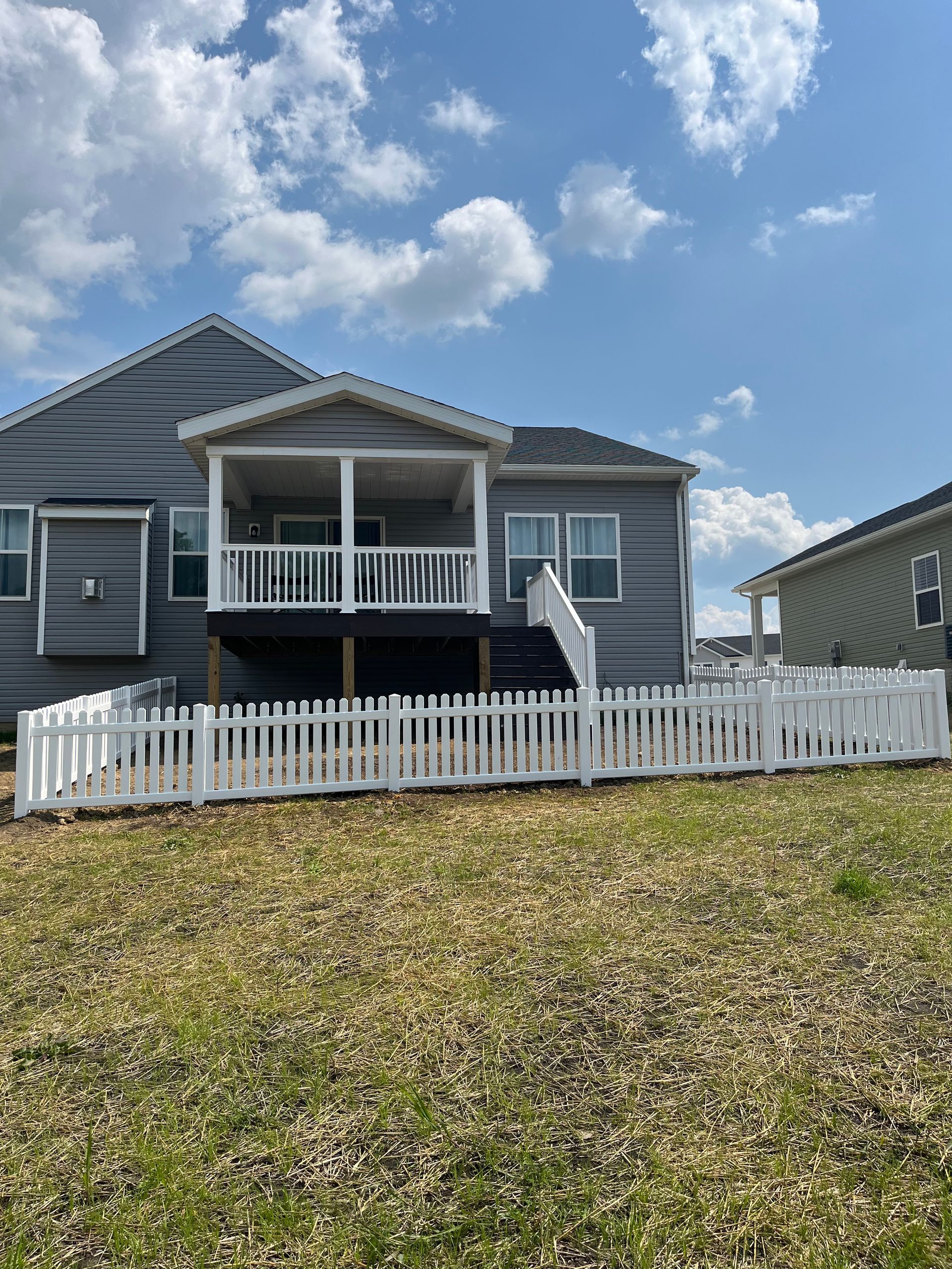Backyard of a gray house with a white picket fence, deck, and covered porch under a blue sky.