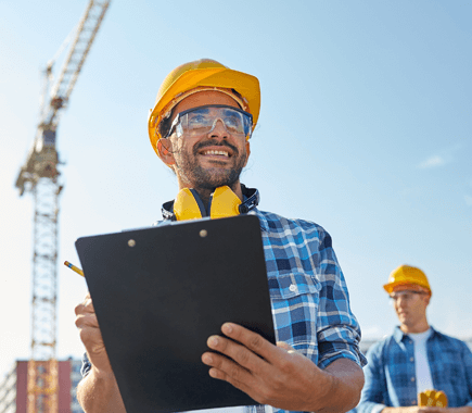 Construction worker in yellow hard hat, holding clipboard, smiling at a construction site with a crane.