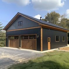 Black metal building with wood accents, garage doors, and a small front door; set on a paved area with greenery.