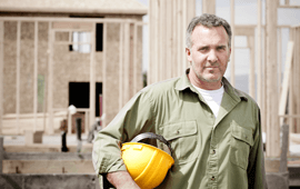 Construction worker holding a yellow hard hat, in front of a wooden house frame under construction.