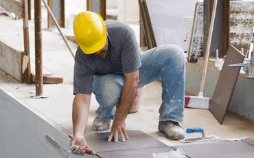 Construction worker laying tile, wearing a yellow hard hat, crouching, holding a trowel, outdoors.
