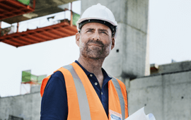 Construction worker in safety vest and hard hat, holding blueprints, looking up, on a construction site.