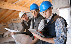 Three construction workers reviewing blueprints and a tablet in an attic, wearing hard hats and vests.