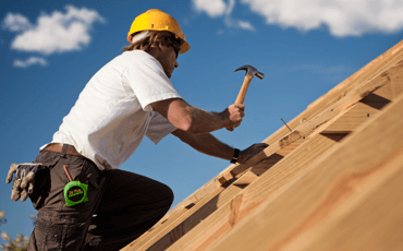 Construction worker in yellow hard hat hammering on a wooden roof under a blue sky.