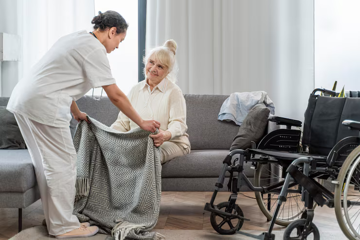 Caregiver assisting an older person with a blanket on a couch, near wheelchairs.