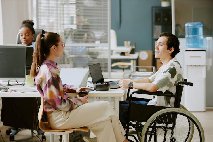 Woman and man in wheelchair talking at a desk, another person at a computer behind them.