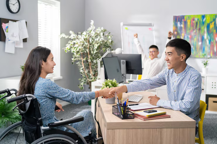 Woman in wheelchair shaking hands with a man at a desk. Another man cheers in the background. Office setting.