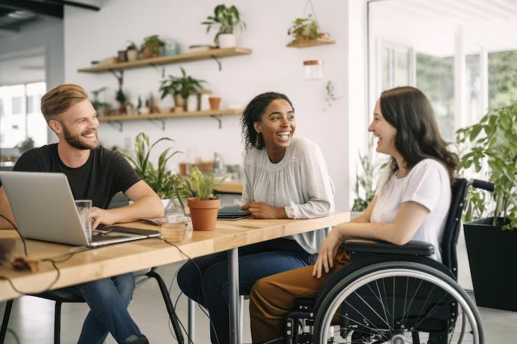 Three people, one in a wheelchair, laughing around a table with a laptop and plants in a bright room.