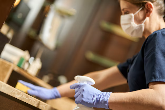 Person in mask and gloves cleans a surface with a spray bottle and sponge.