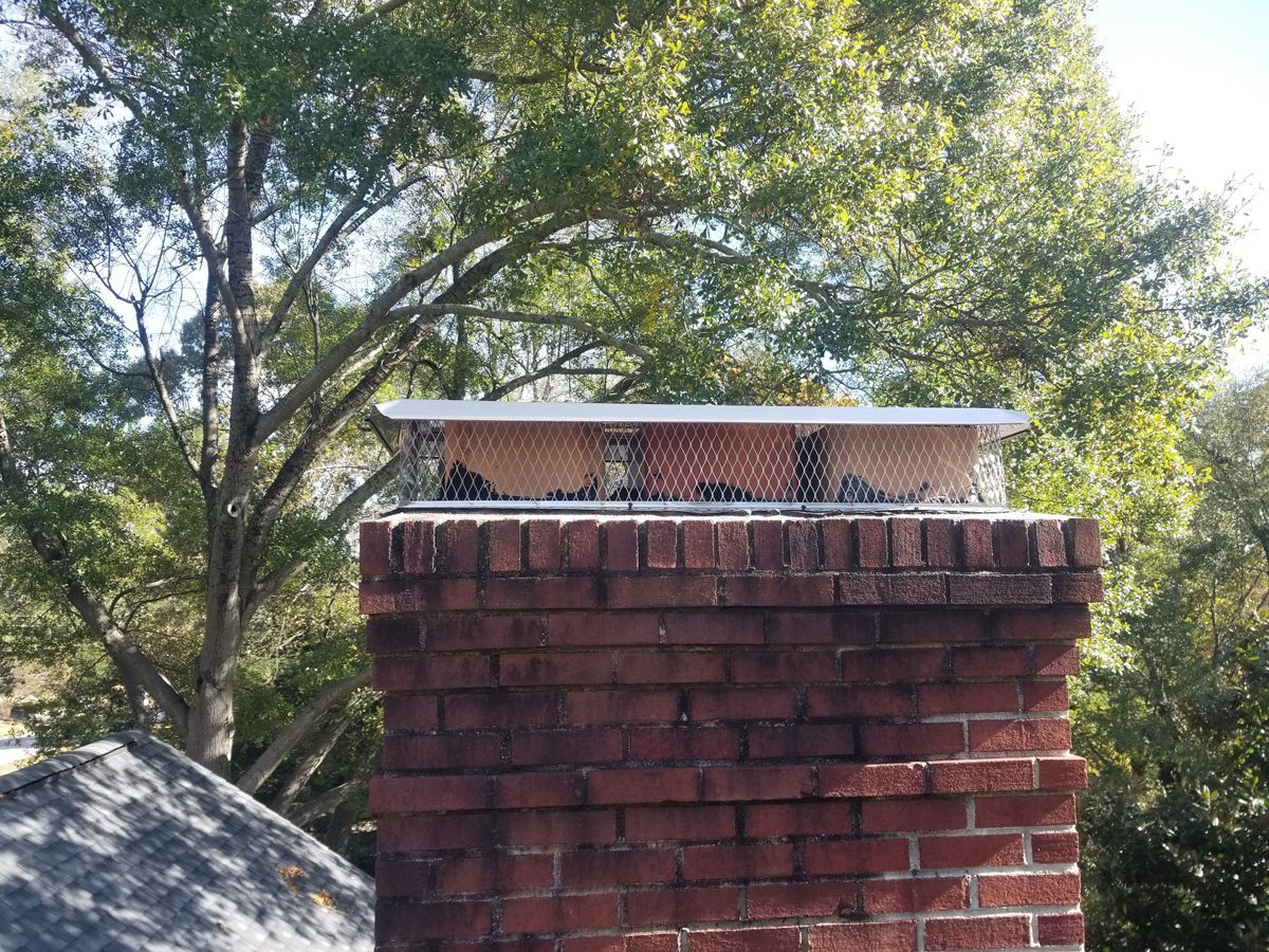 Brick chimney with a metal mesh cap, set against a background of trees.