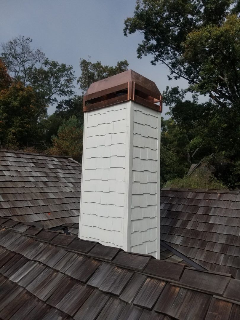 Copper-topped chimney on a shingled roof, surrounded by trees under a cloudy sky.